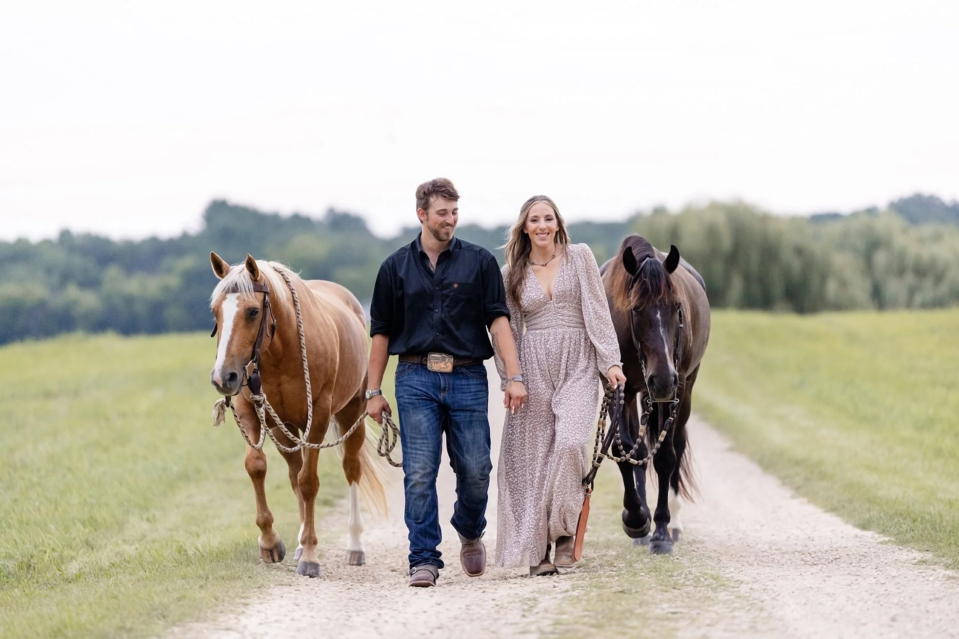 Amber and Isaac walk hand in hand down a gravel farm road with their horses, Atlas and Ellie, during their western engagement session at Advanced Horsemanship in Poynette, Wisconsin.