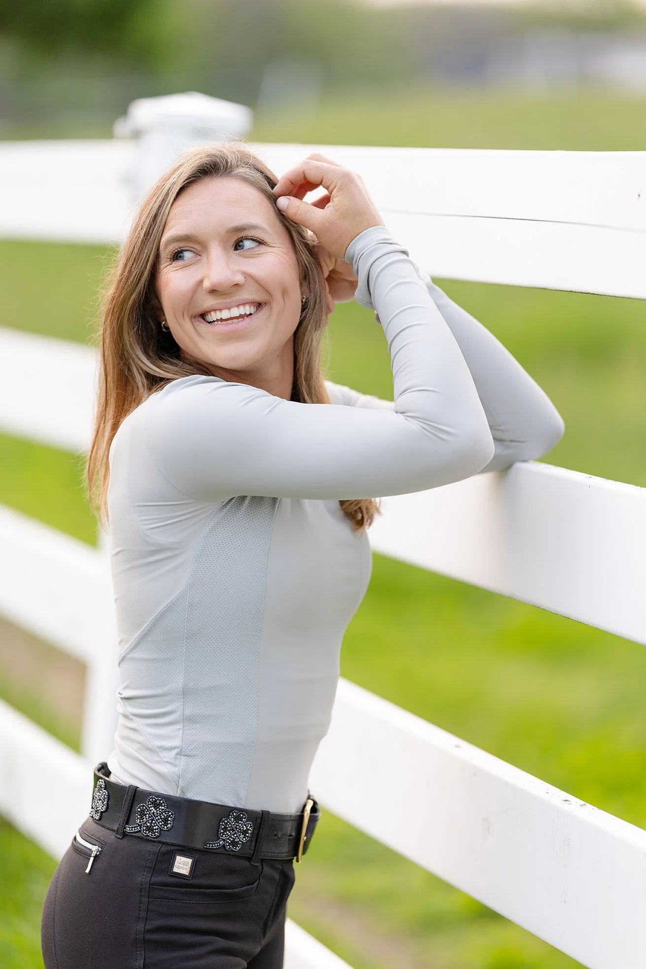 Smiling equestrian rider in STS Equestrian light grey shirt leaning against white fence.