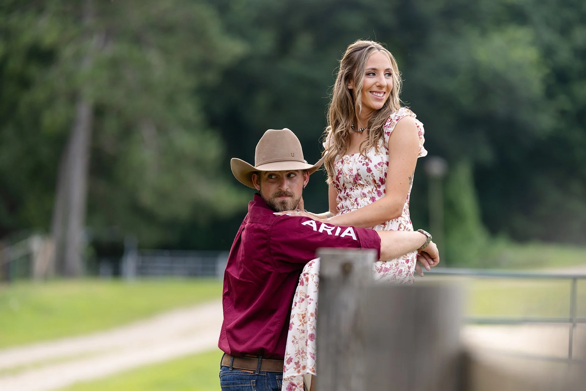 Amber smiles while Isaac lifts her beside a wooden fence during their western engagement session in Poynette, Wisconsin. A soft, natural background highlights the romantic setting on their horse training property.