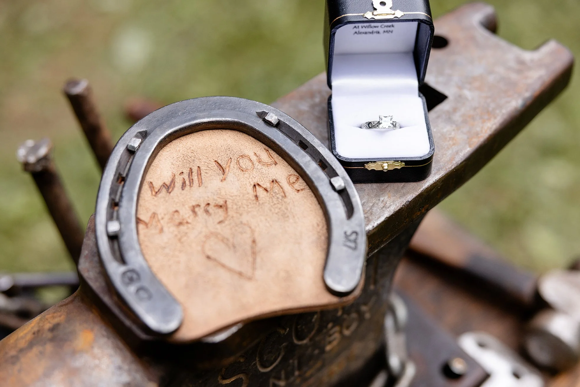 A carved leather horseshoe reading “Will you marry me?” sits beside Amber’s engagement ring box on an anvil, capturing a meaningful western-inspired proposal detail during their engagement session.