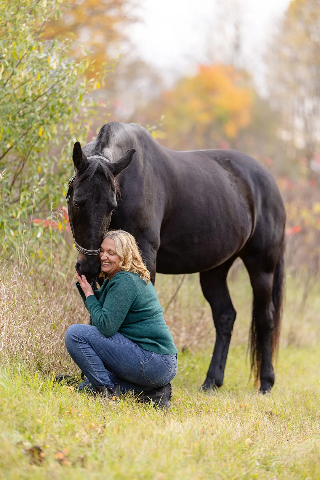 Julie kneels beside her dressage horse during a fall equine portrait session in Plymouth, Wisconsin, framed by tall grasses and fall foliage.