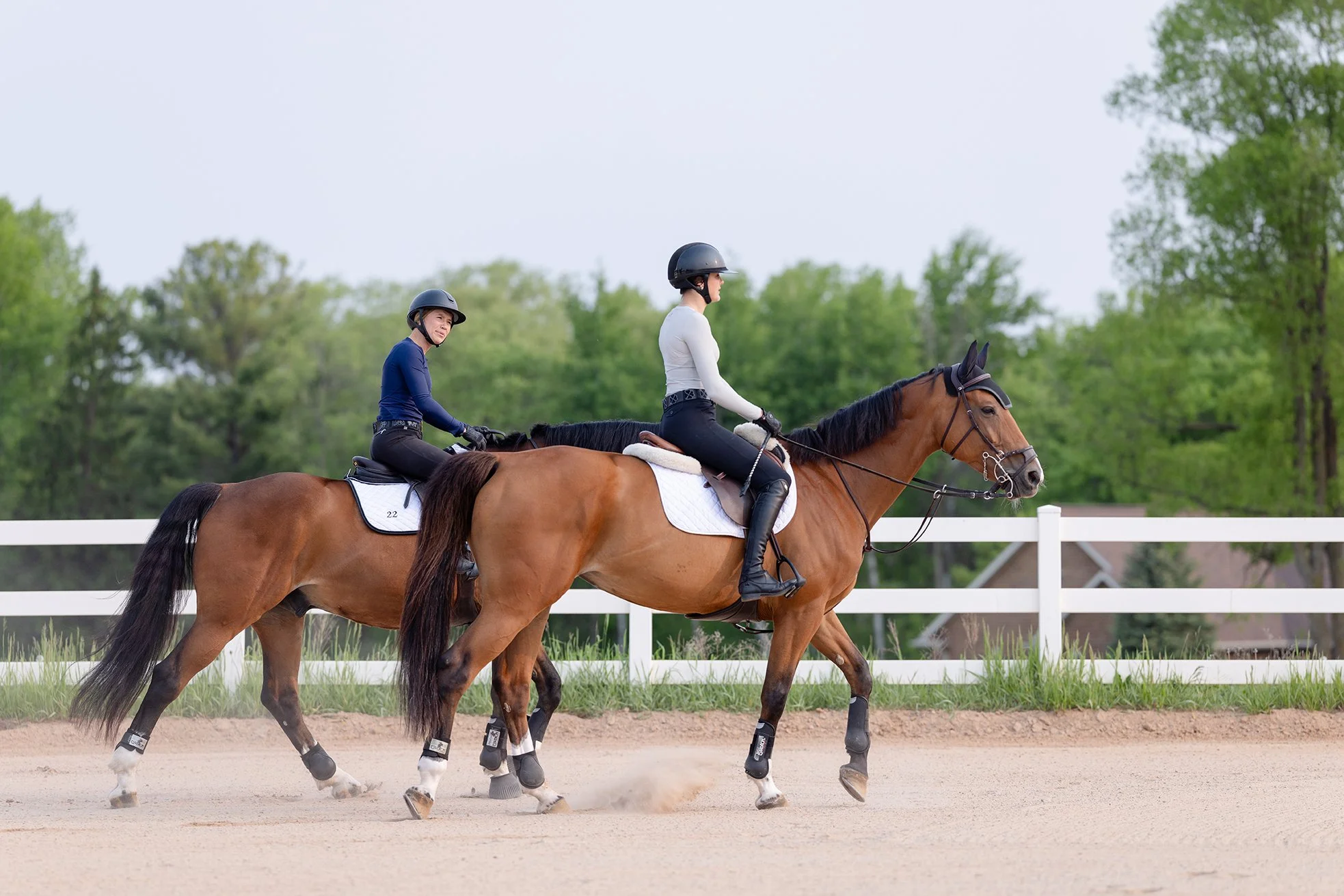 Two riders in STS shirts riding side by side in a sand arena with white fencing.
