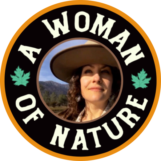 Person wearing a wide-brimmed hat and overalls, smiling and holding a piece of hair or beard under their nose, standing outdoors with trees in the background.