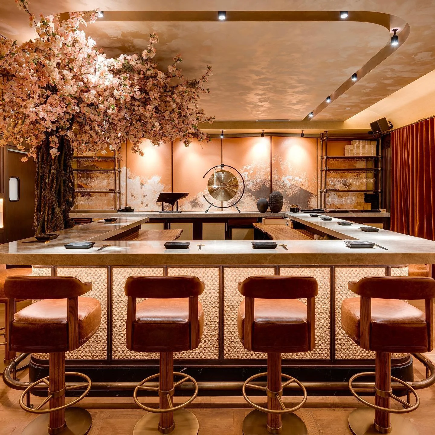 Elegant restaurant interior with a cherry blossom tree decoration on the left, a marble counter with four bar stools in the foreground, and a decorative wall with a gong, vases, and shelving in the background.