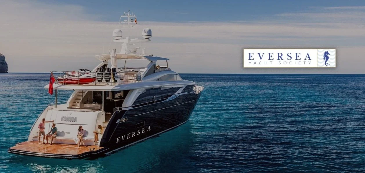 Luxurious black yacht named Eversea anchored in clear blue waters near a rocky coastline under a partly cloudy sky, with four people sitting on the deck and a sign in the background reading Eversea Yacht Society.