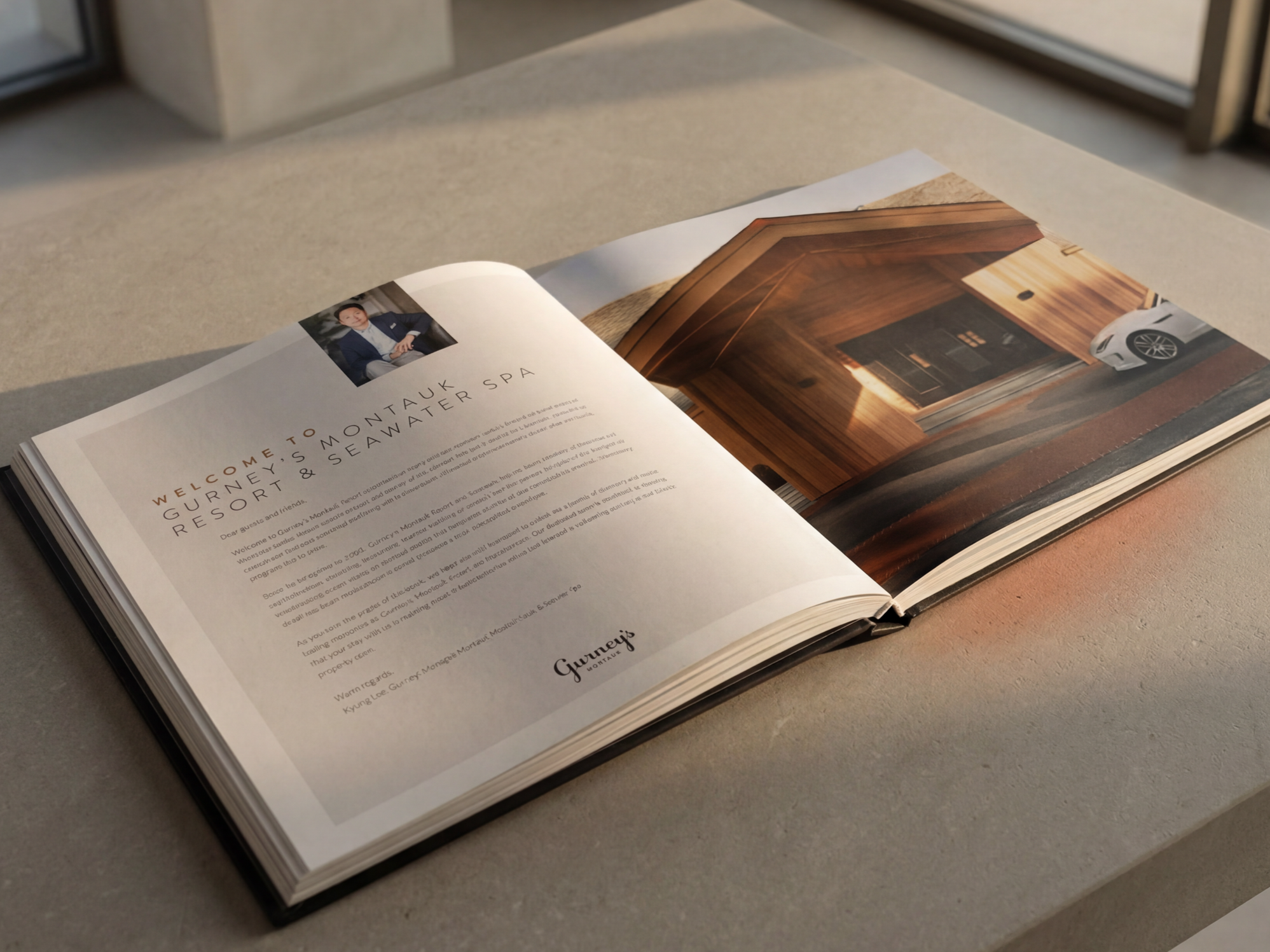Open hardcover book on a table displaying a page with a photo of a man in a suit, text about Gurney's Montauk Resort & Seawater Spa, and a large image of a wooden house exterior with a white car parked outside.