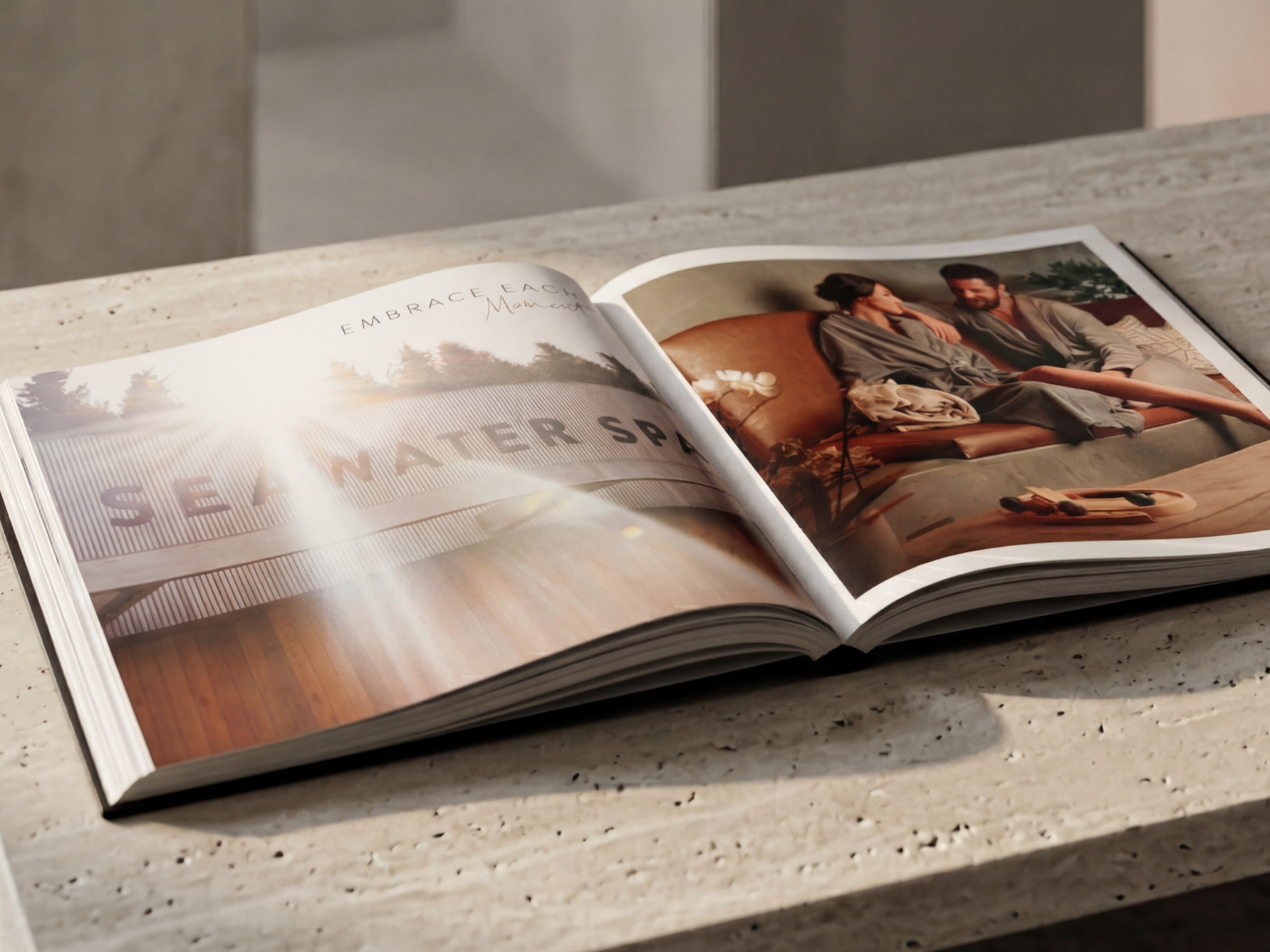 Open hardcover book on a marble table showing two pages, one with a title about embracing each moment and a photo of a modern building, the other with a photo of a man and woman sitting on a leather couch, talking.