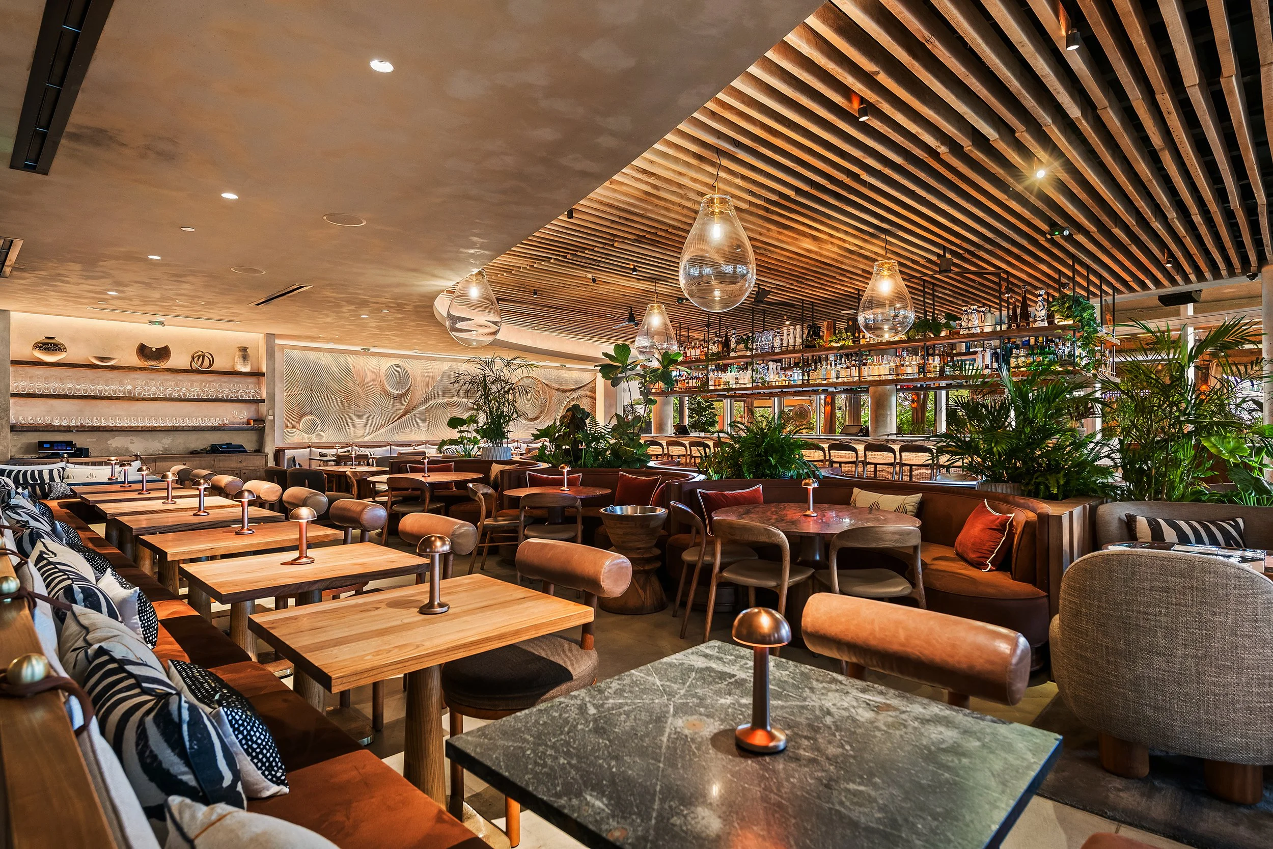 Interior of Mila, a modern restaurant with wooden furniture, decorative vases on shelves, hanging glass light fixtures, and plants near the bar area.