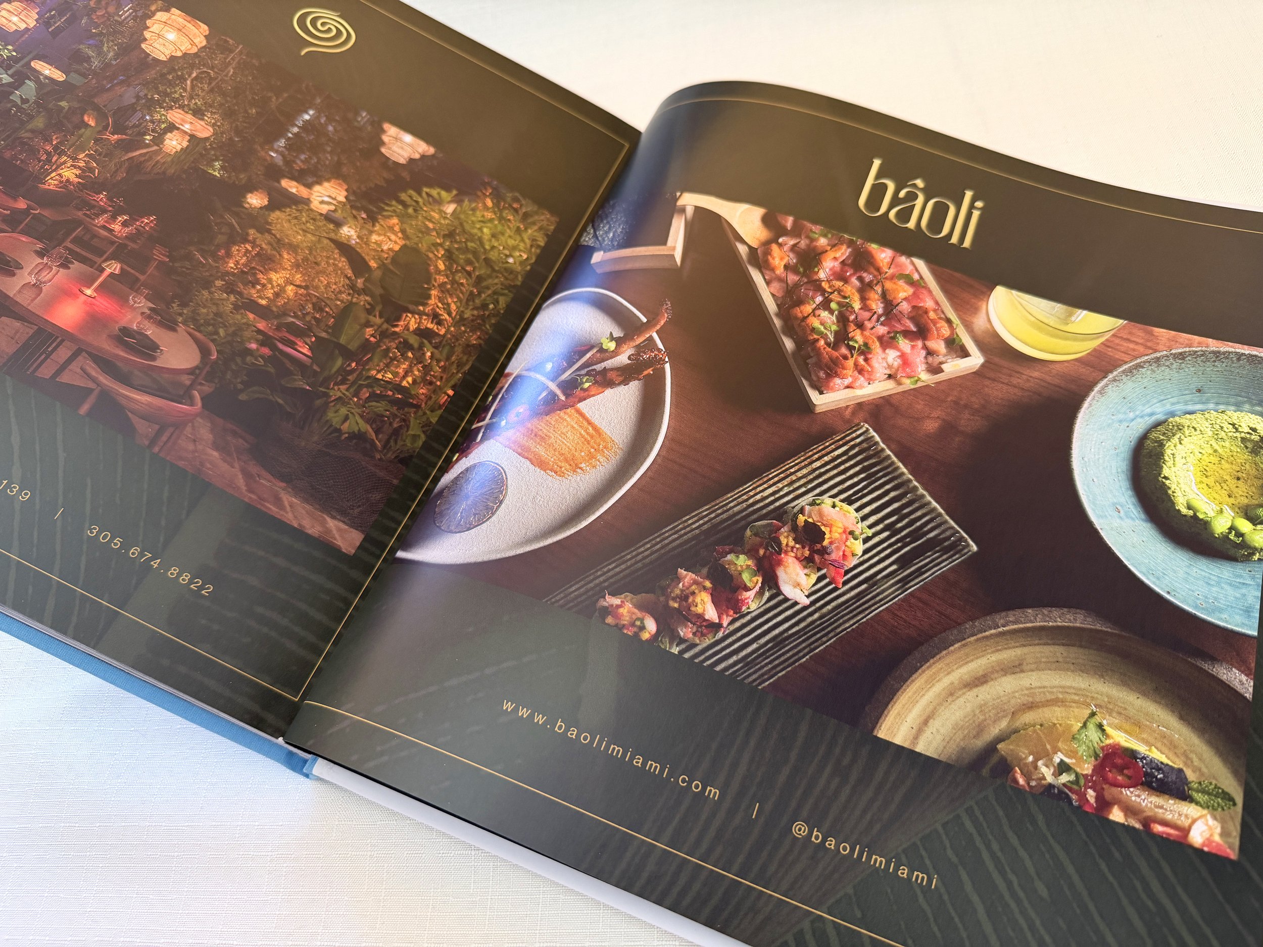 Open magazine showing colorful images of Asian cuisine including sushi, rolls, and salads, with the website URL www.baolimiami.com visible.