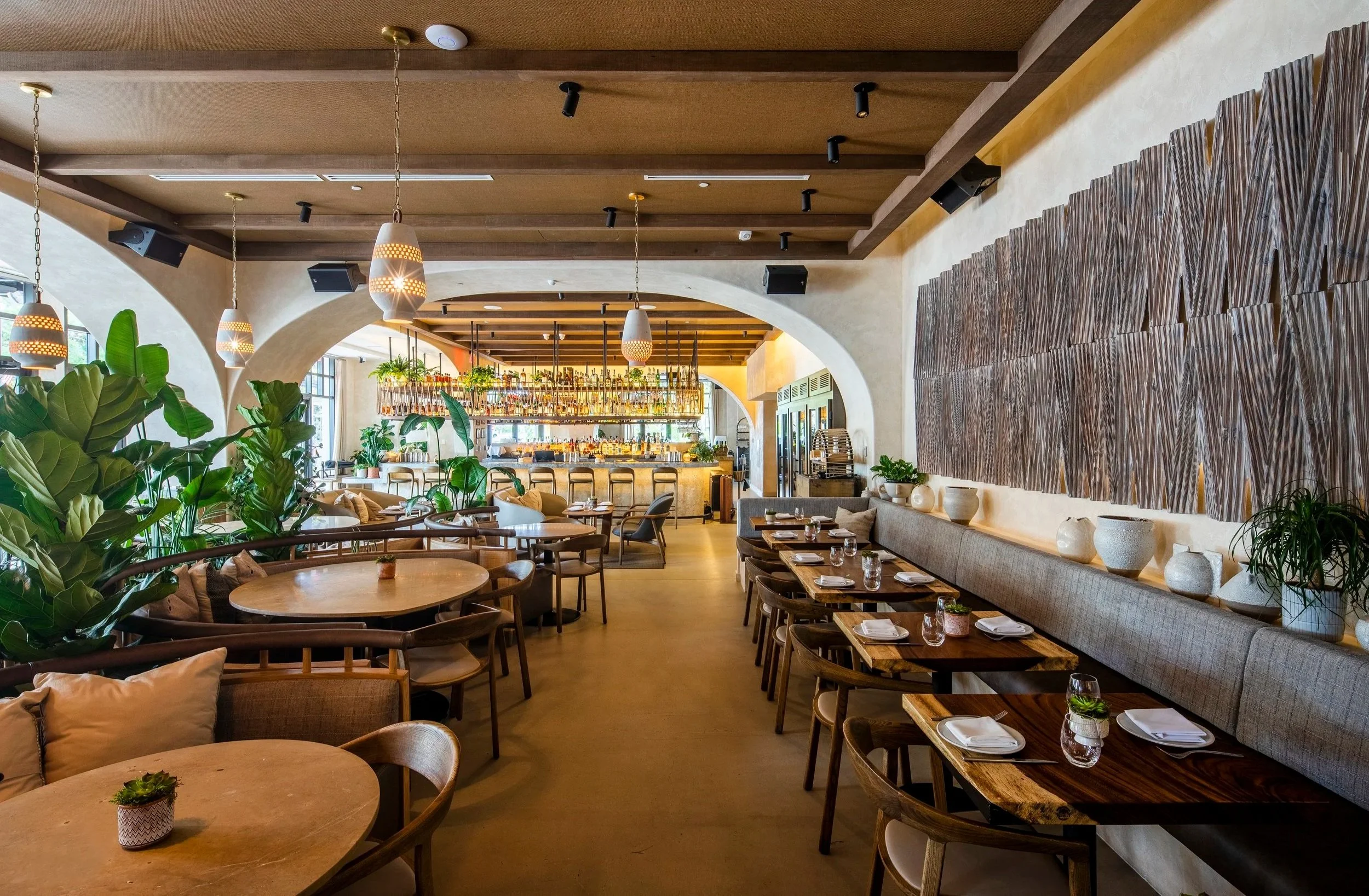 Interior of a modern restaurant with wooden furniture, potted plants, decorative vases, pendant lighting, and a bar area in the background.