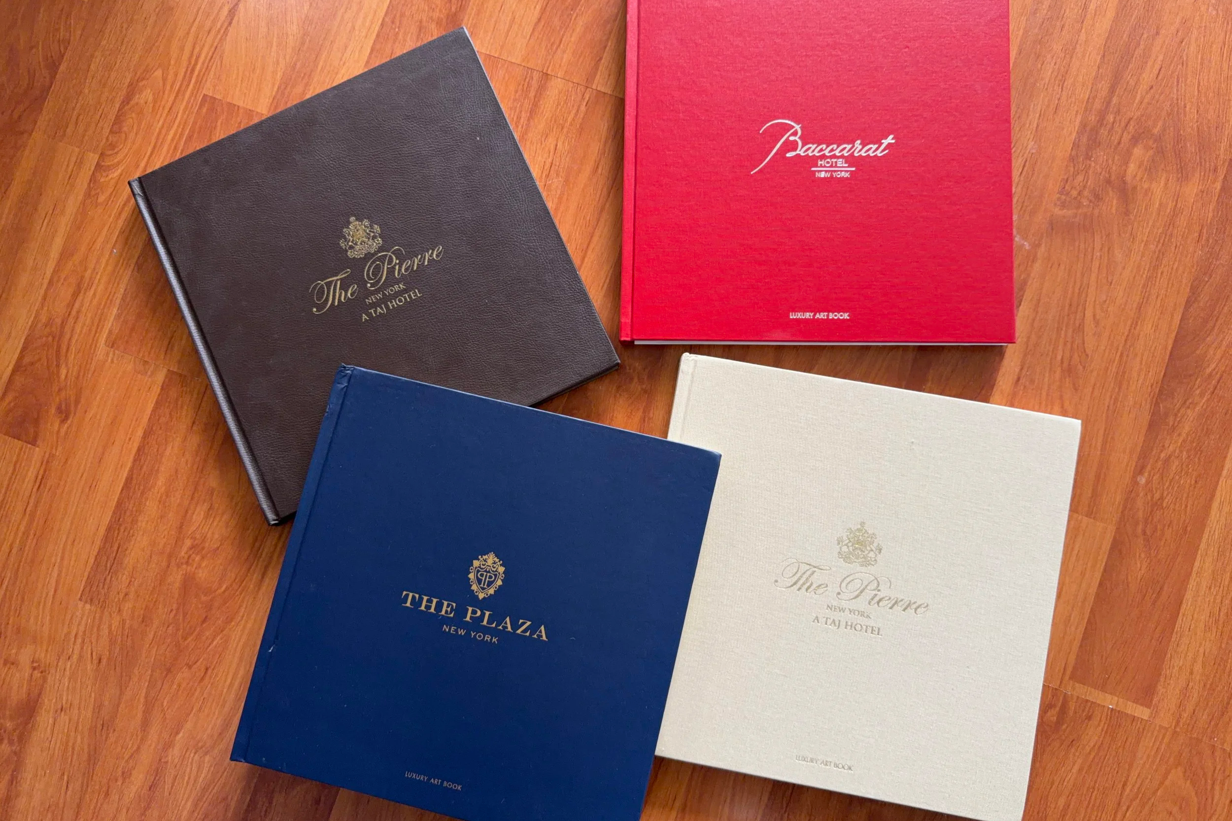 Four hardcover books on a wooden floor, two with gold embossed text and logos, titled 'The Pierre' and 'The Plaza', and two with a red and white cover, labeled 'Baccarat' and 'Luxury Art Book'.