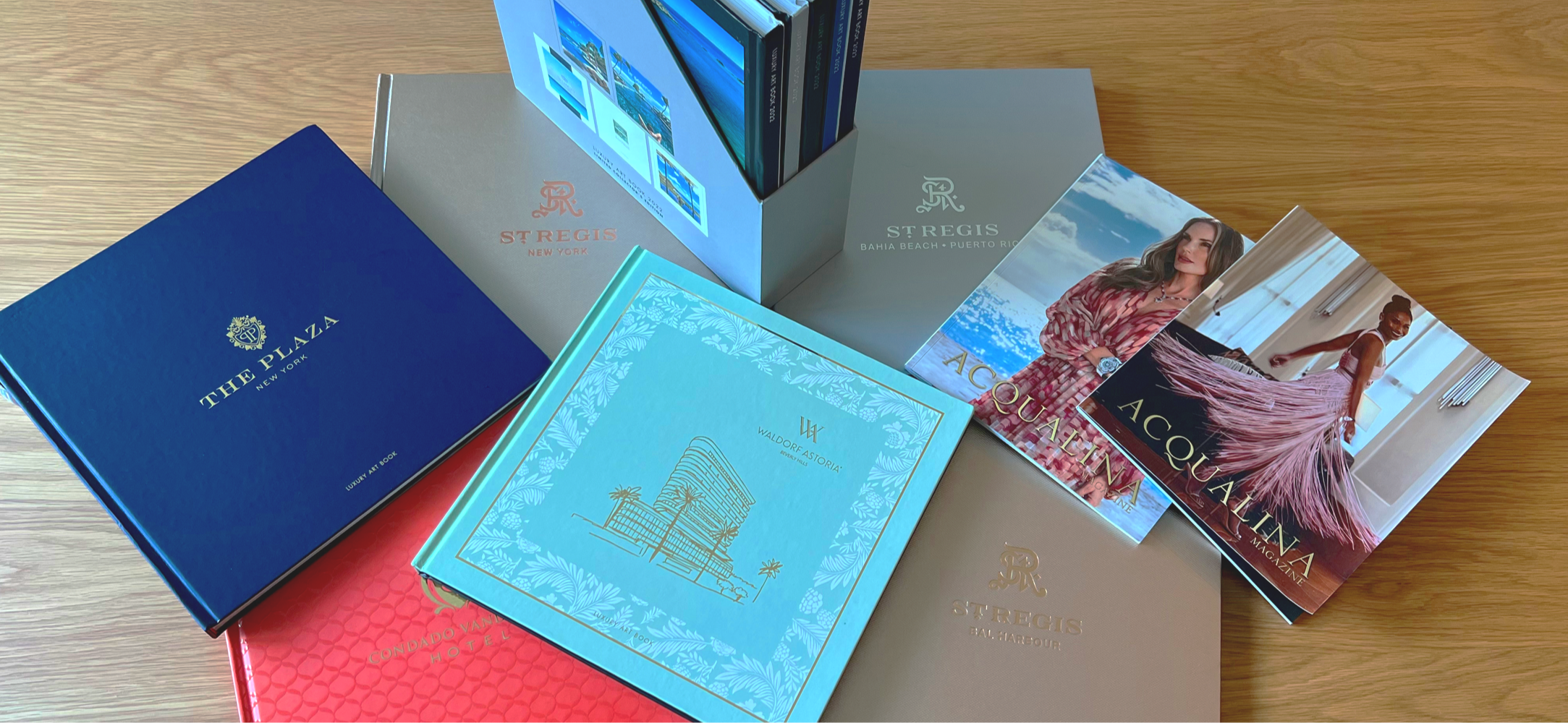 A collection of colorful hotel brochures and magazines on a wooden surface, including The Plaza, Waldorf Astoria, Condado Vantage Hotel, St. Regis in New York, Bahía Beach in Puerto Rico, and magazines featuring fashion models.