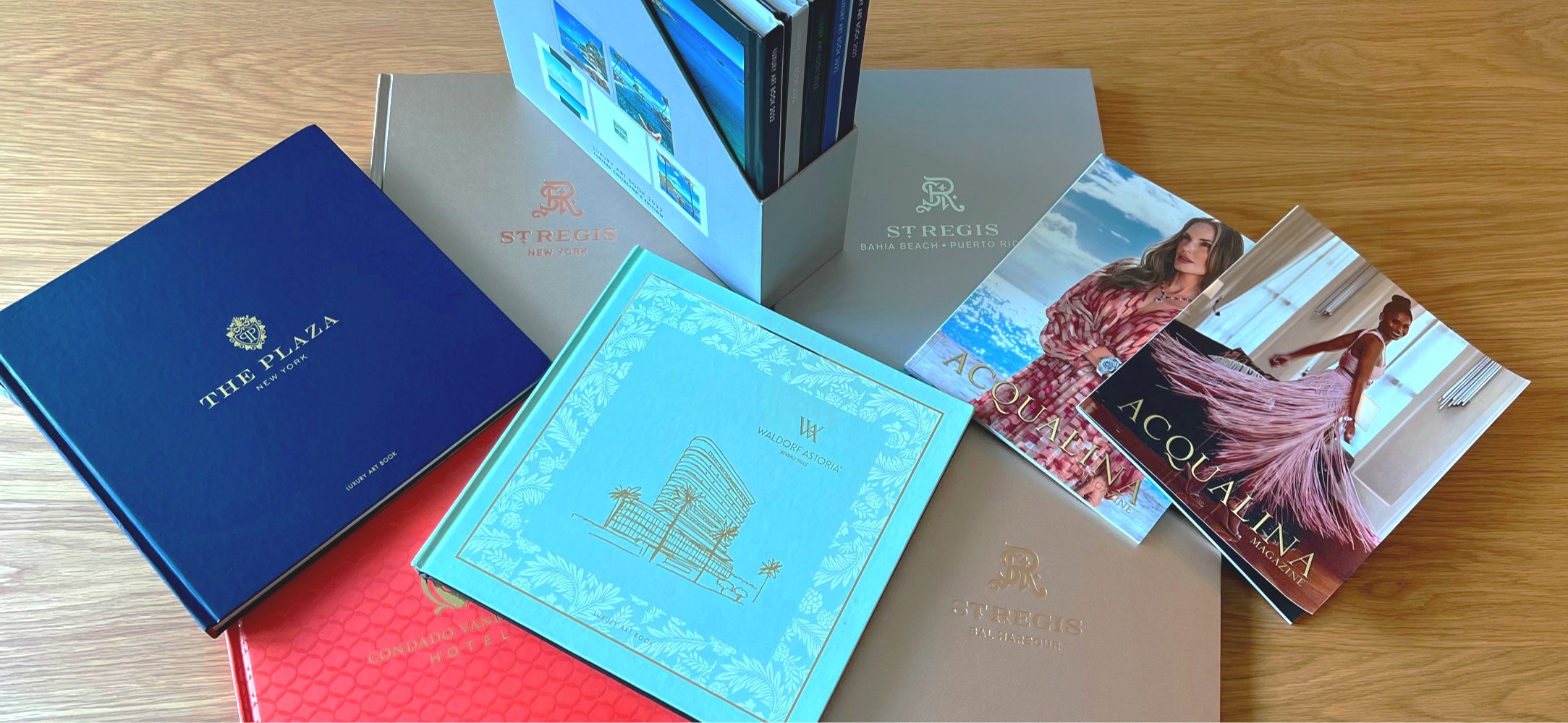 A collection of printed brochures and booklets for luxury hotels including The Plaza New York, Waldorf Astoria Amelia Island, Condado Vanda Hotel, St. Regis properties in New York, Bahia Beach, and Puerto Rico, as well as Acqualina Magazine, spread out on a wooden surface.