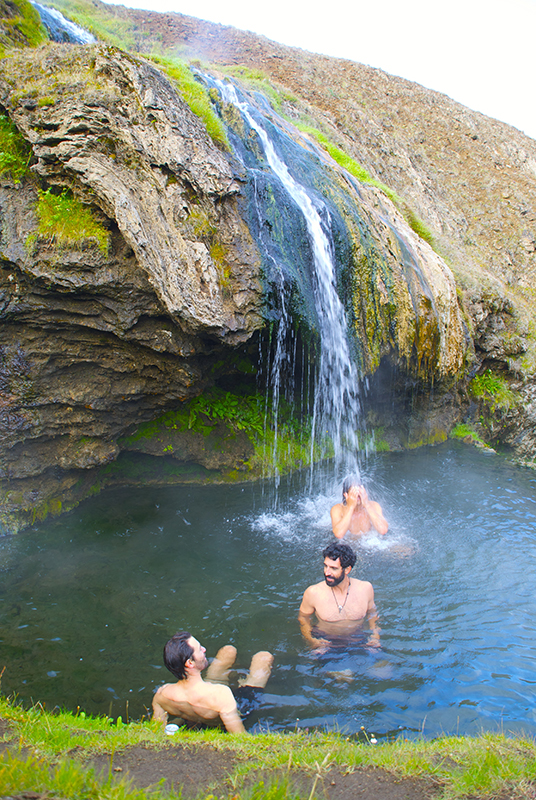Travelers soaking in a natural hot spring waterfall on an Ashram Iceland journey, a unique treat while hiking in Iceland.