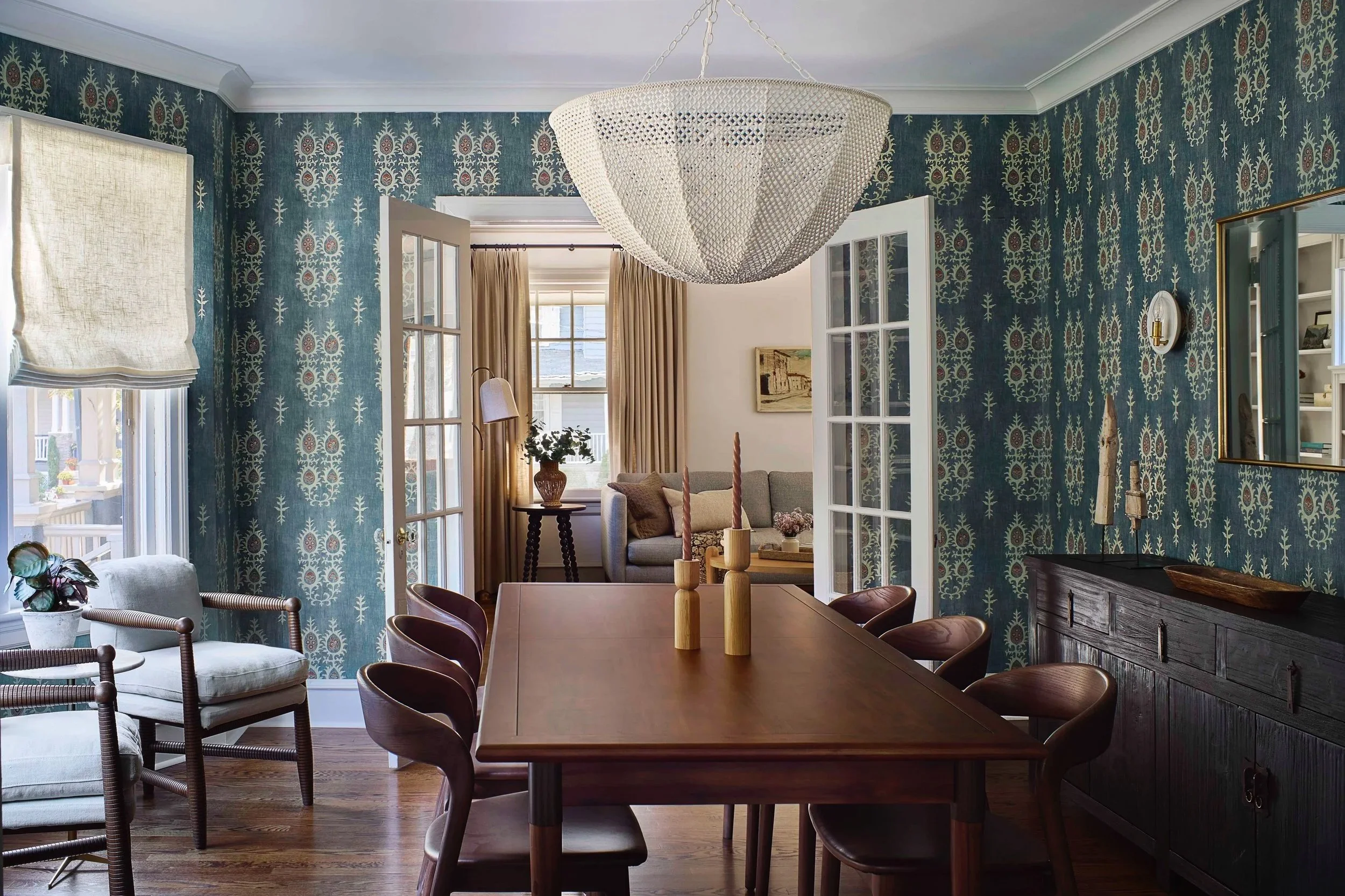 Elegant dining room with Moroccan inspired patterned wallpaper, wood dining table and chairs, woven chandelier, and French doors opening into a cozy sitting room.