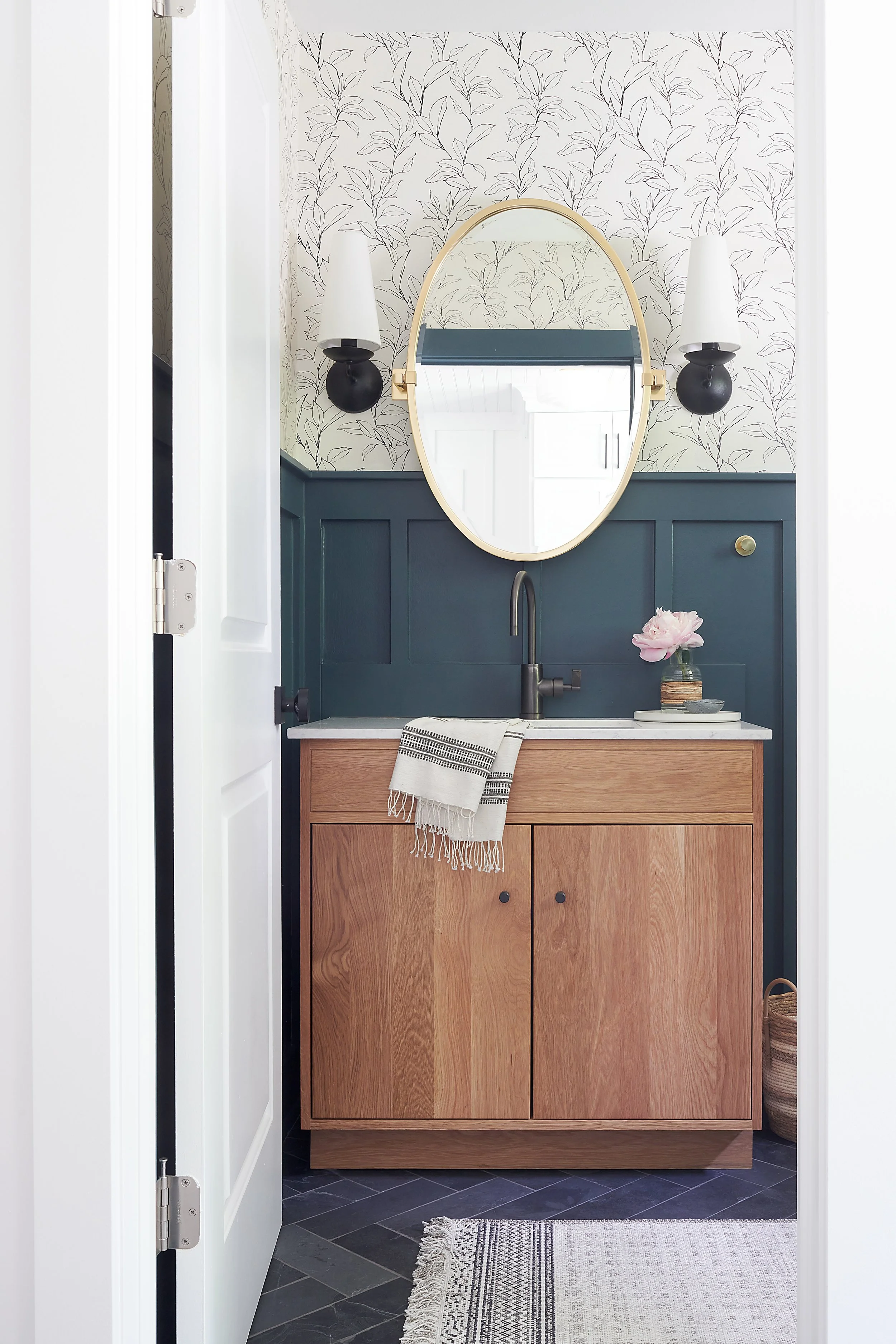 Powder bathroom design with natural stone herringbone pattern floor tile, blue wainscot millwork, white and black botanical print wallpaper