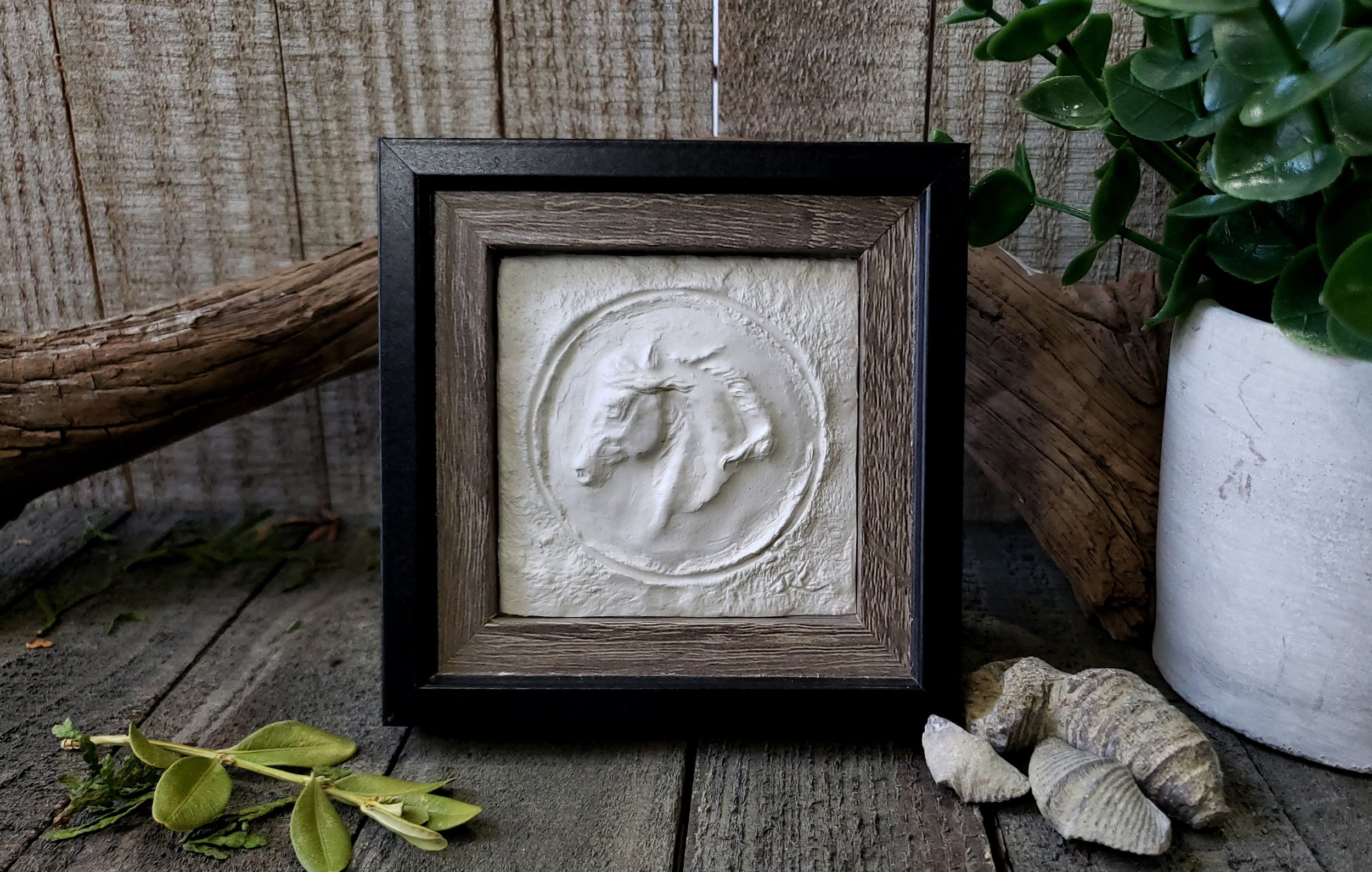 sculpture small 4x4 white horse framed in black sitting on wood background with plant beside.jpg