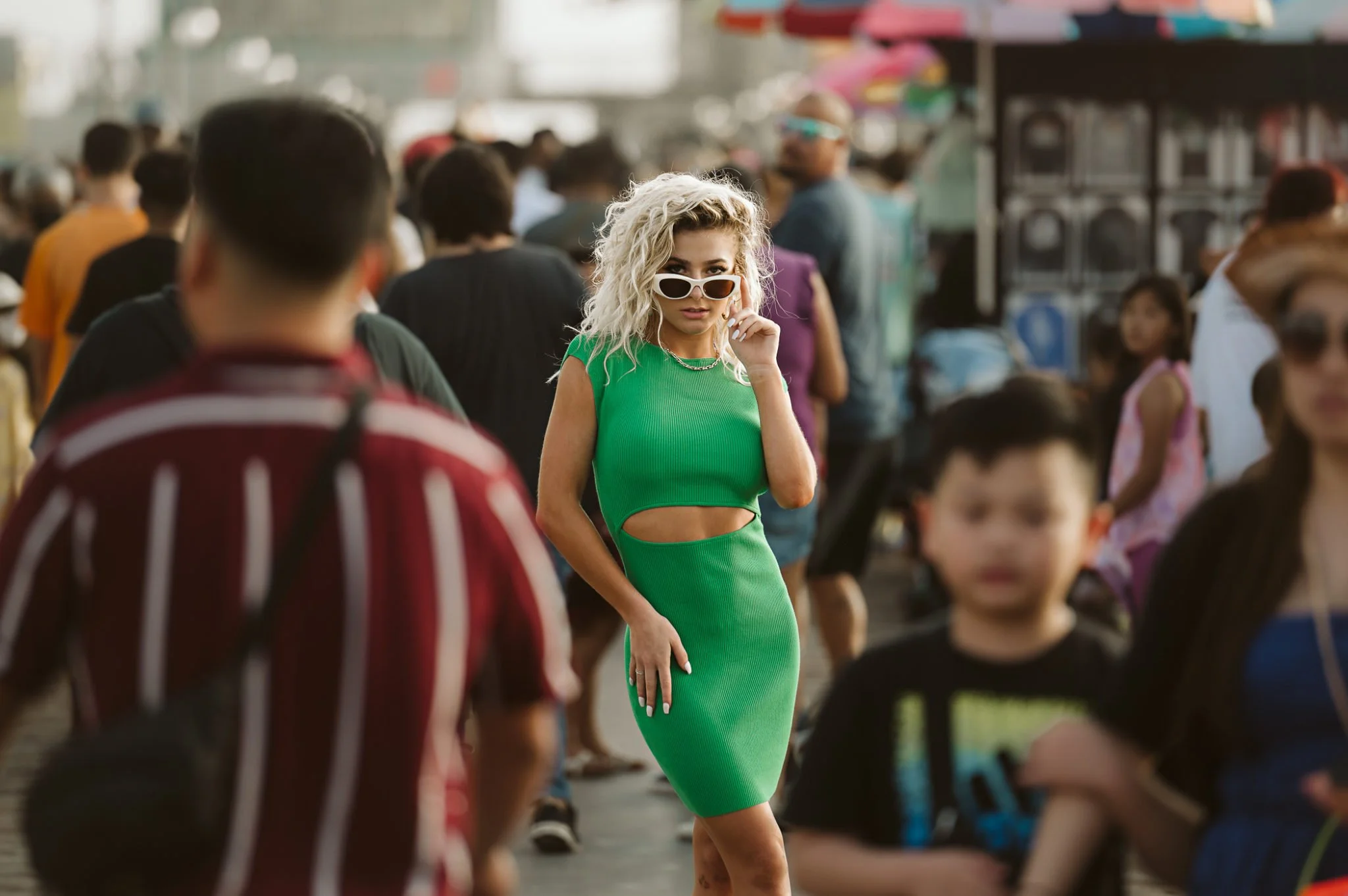 A woman with curly blonde hair wearing a fitted green dress and white sunglasses stands amidst a crowd of people, some blurred, in an outdoor marketplace or street.