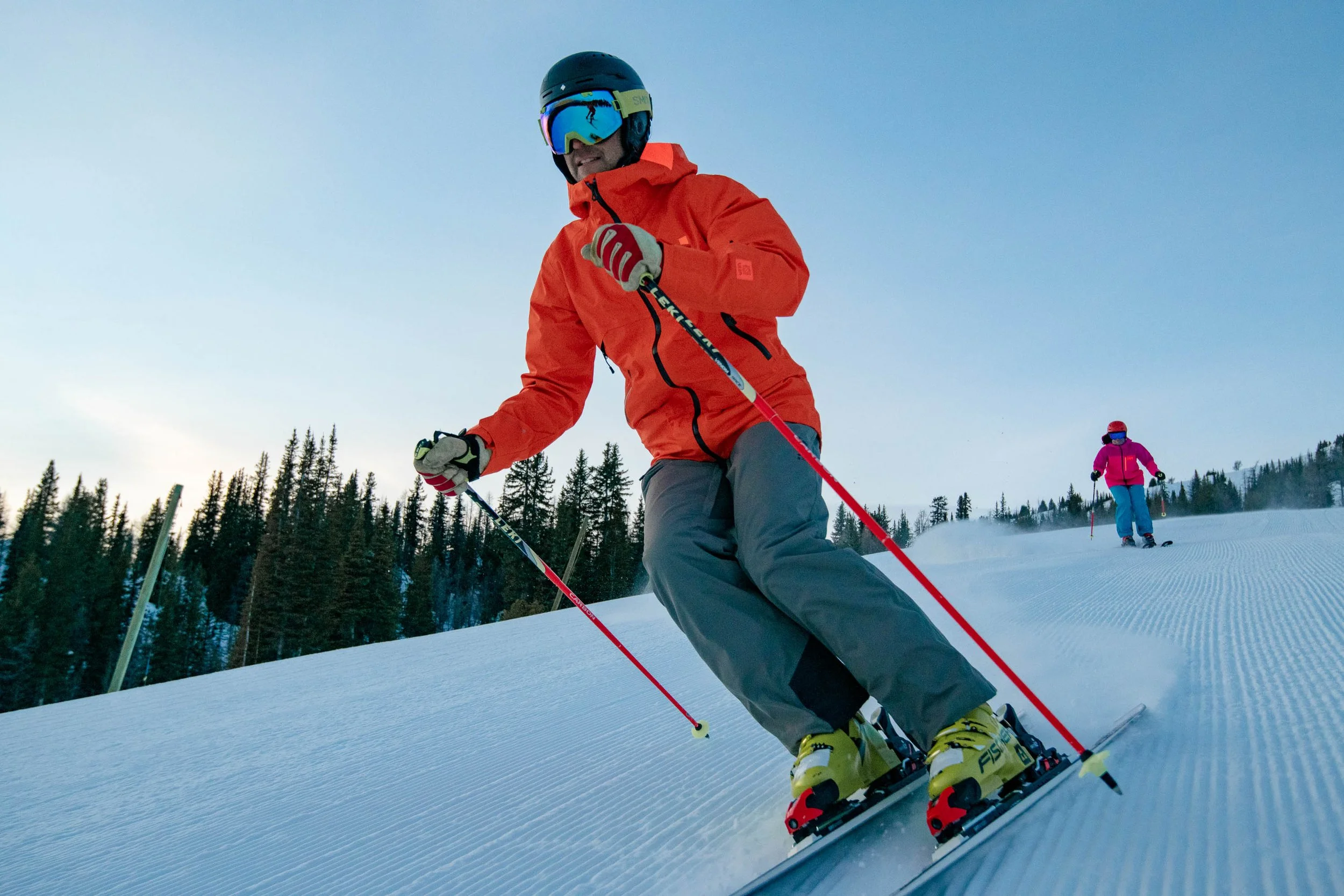A man in an orange jacket, gray ski pants, yellow skis, and yellow ski boots skiing downhill on a snow-covered slope, wearing a black helmet, reflective ski goggles, and glasses. In the background, a woman in a pink jacket and blue pants skis further down the slope, with a forest of pine trees and a cloudy sky visible.
