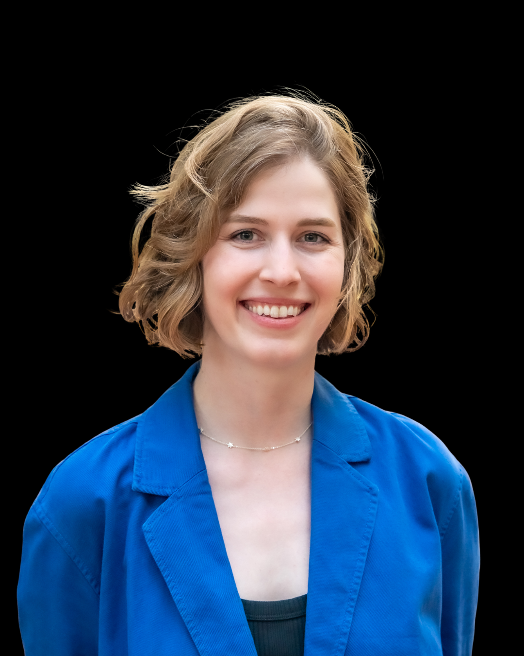 Smiling young woman with short, wavy blonde hair, wearing a blue blazer and a necklace, against a black background.