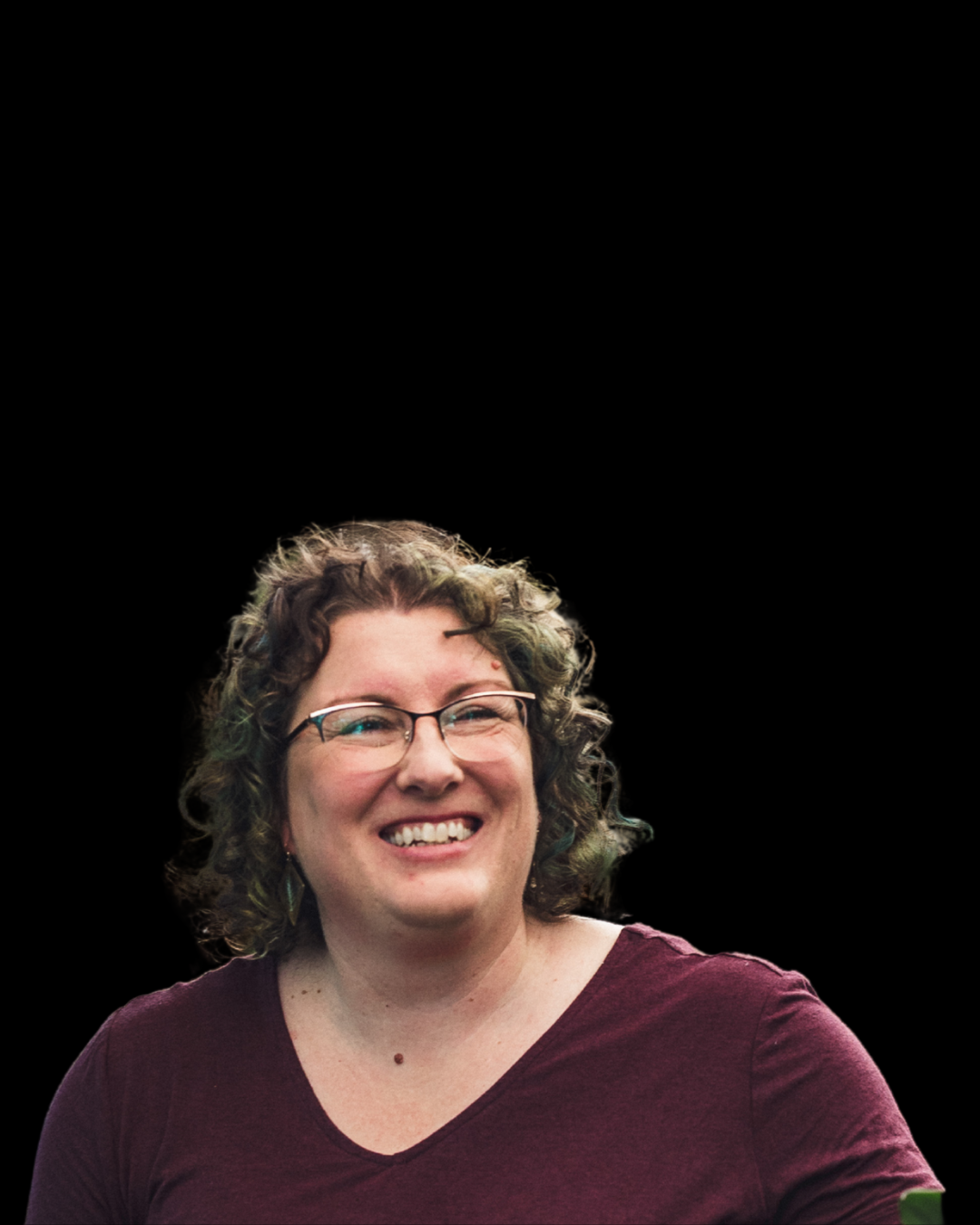A smiling woman with curly hair, glasses, and a maroon shirt against a black background.