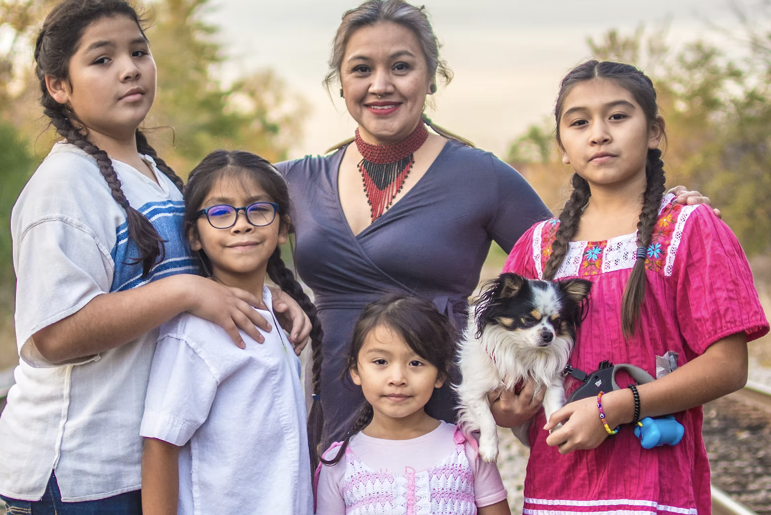 A smiling woman surrounded by four young girls and a small dog outdoors, with trees and sky in the background.