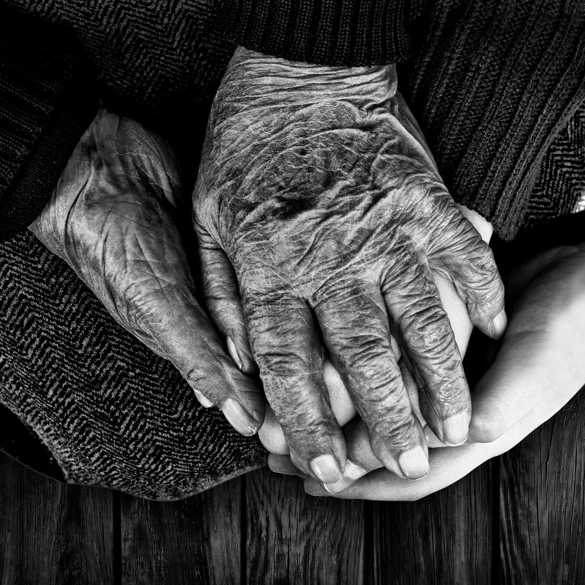 Close-up of elderly person's wrinkled hand gently holding a younger person's hand, both resting on a wooden surface.