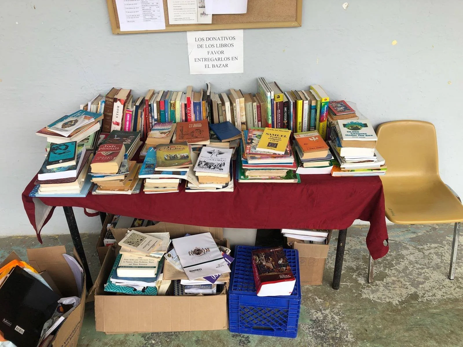 A table covered with a red cloth filled with stacked books for donation or sale, with boxes and crates underneath holding more books, set against a plain wall with a bulletin board above.