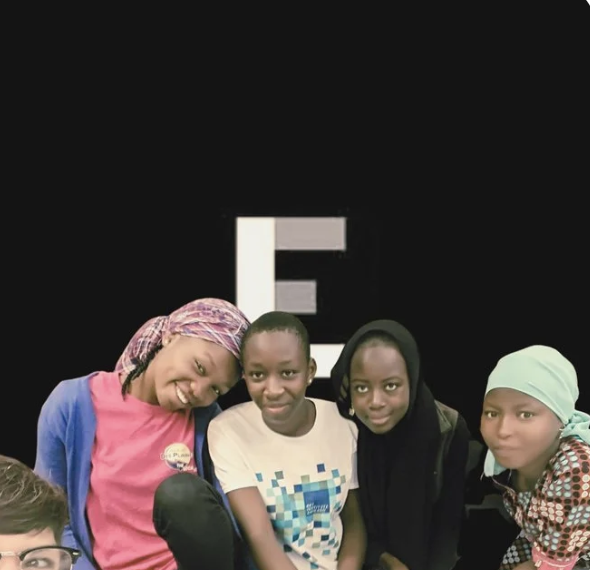 Four young girls sitting together in front of a black background with a large letter 'E' behind them for Expanding Lives nonprofit logo representing community support, empowerment, and holistic wellbeing
