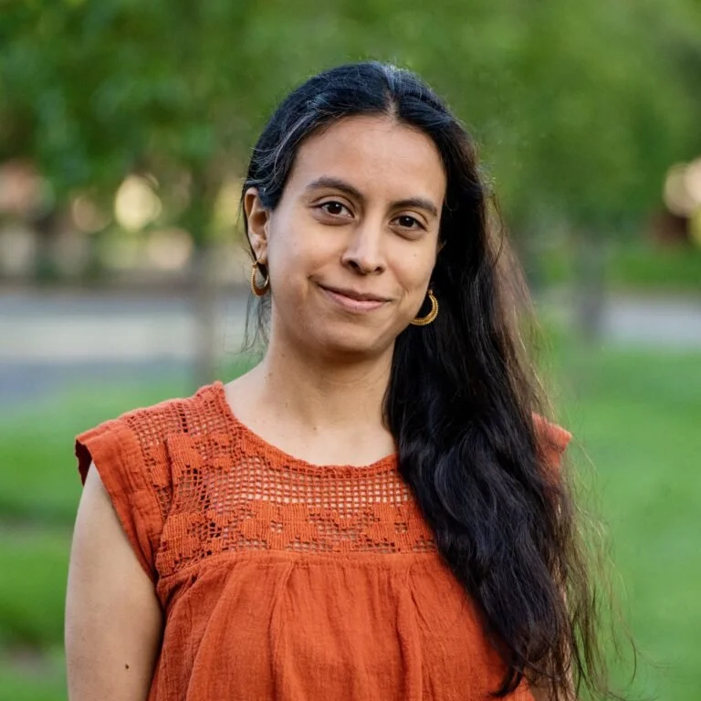 A woman with long dark hair and hoop earrings smiling outdoors in a park, wearing an orange sleeveless top with crochet details.