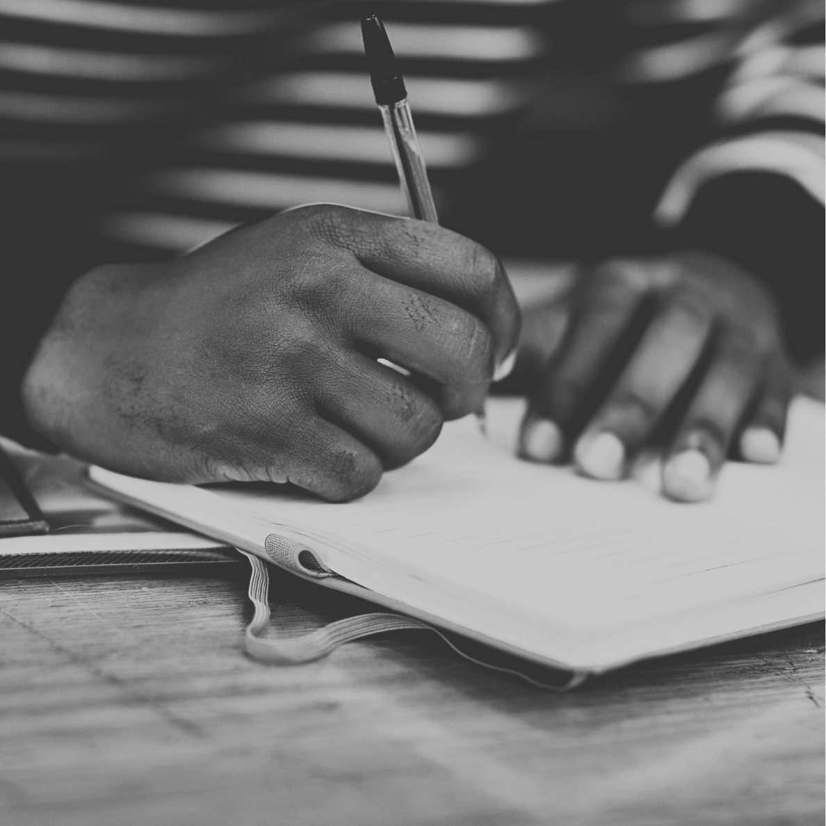 A black-and-white close-up of a person's hand holding a pen and writing in a lined notebook on a wooden surface.