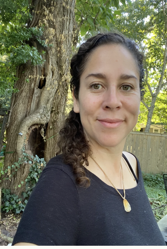 A woman with curly dark hair pulled back, wearing a black shirt and a gold necklace with a pendant, standing outdoors in front of a large tree and a wooden fence, smiling at the camera.