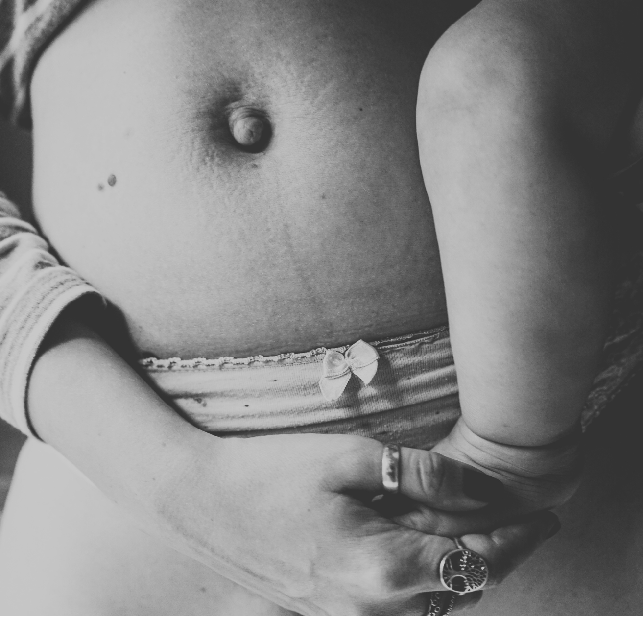 Close-up of a pregnant woman's belly with hands gently resting on her stomach, wearing rings and a striped shirt with a small bow detail.