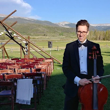 A man dressed in a tuxedo holding a cello outdoors, with chairs and a wooden structure in a field with mountains in the background.