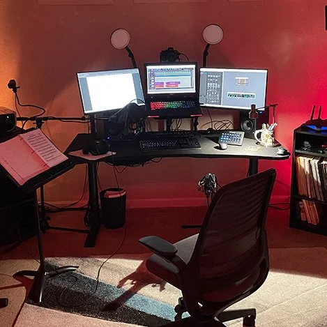 Workspace with a desk holding three monitors, a keyboard, a mouse, and a talk mic, surrounded by various electronic equipment and books, against a red wall.