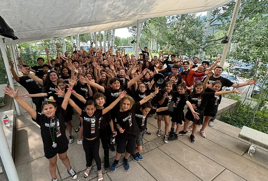 Large group of children and adults posing joyful with raised hands outdoors under a canopy with trees in the background
