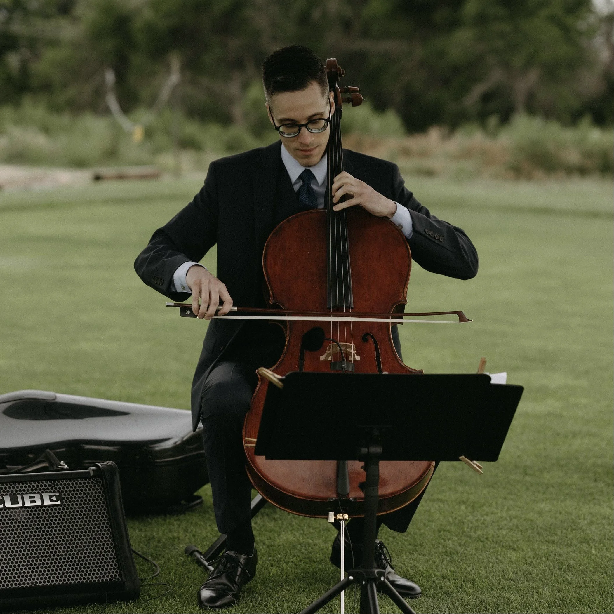 Michael Linert in a suit playing a cello outdoors on a grassy area with trees in the background.