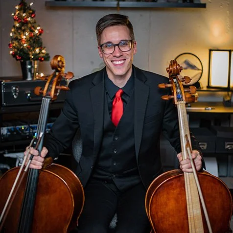 A man in a black suit, red tie, and glasses sitting between two cellos in a recording studio decorated for Christmas.