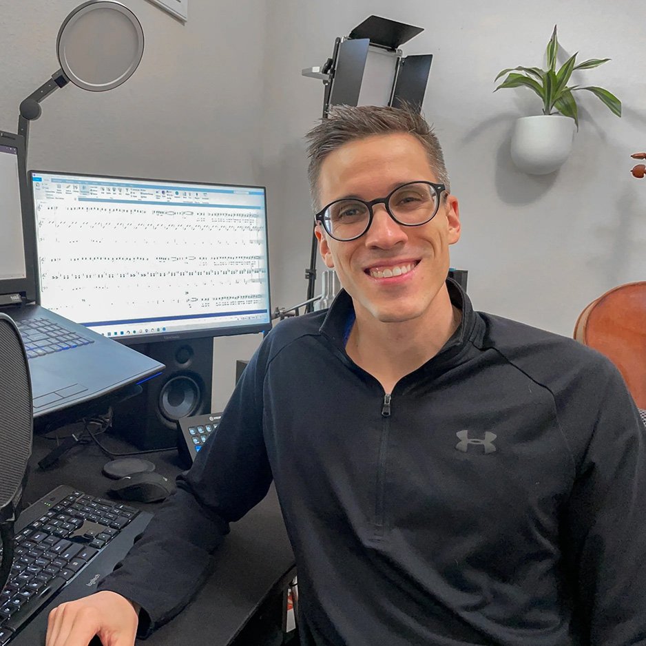 A smiling man wearing glasses and a black jacket sitting at a desk with music production equipment, including a computer displaying sheet music, studio monitor speakers, and a microphone in a home studio setting.