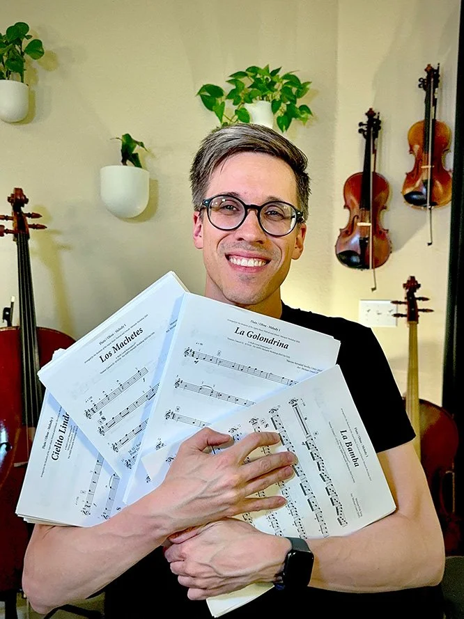 A man smiling and holding sheet music in a room with wall-mounted violins and potted plants.