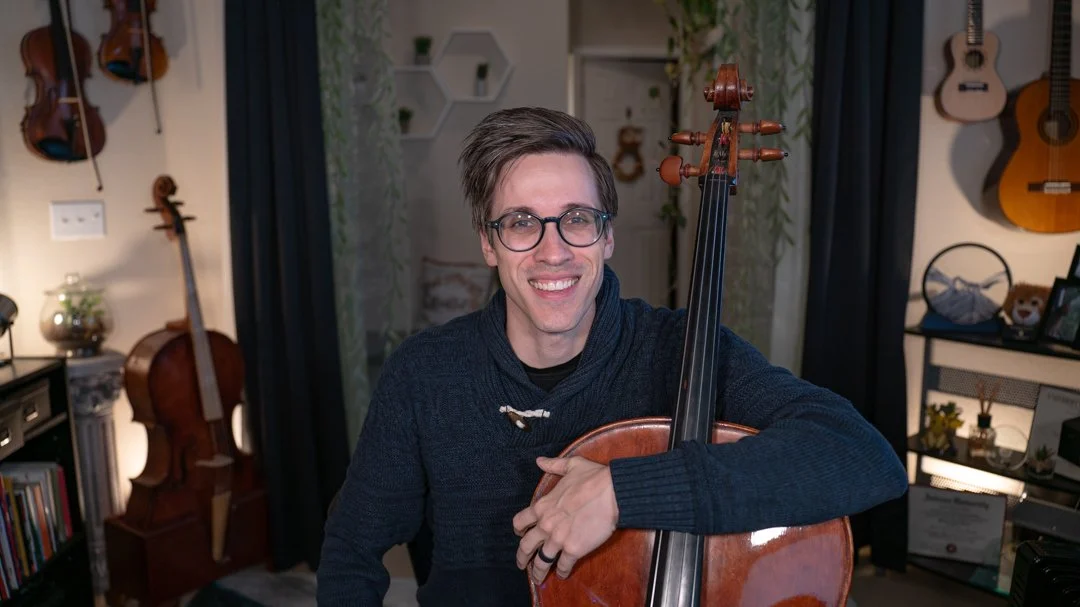 Man with glasses smiling while holding a cello in a room with guitars and musical decor.