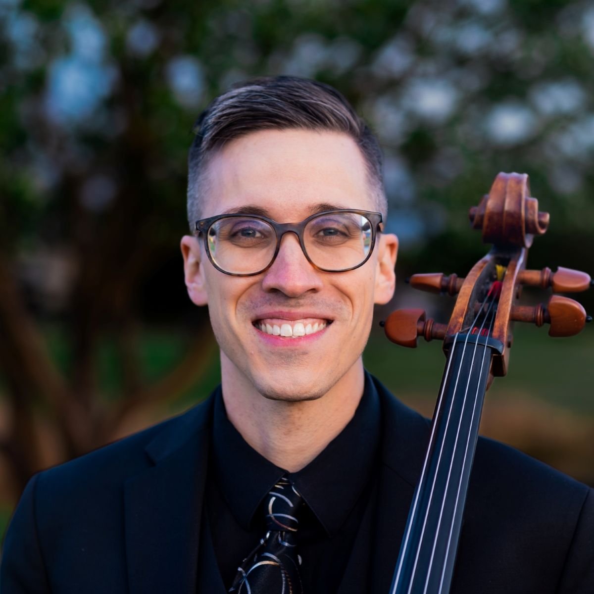 Michael Linert wearing a black suit and striped tie, smiling while holding a cello outdoors with trees in the background.