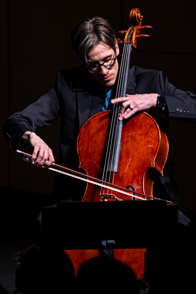 A man in a dark suit and glasses playing a cello on stage.