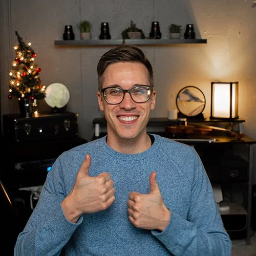 A young man in glasses and a blue sweater smiling and giving two thumbs up in a decorated room with Christmas decorations and modern lighting.