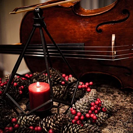 Close-up of a cello, red candle inside a geometric candle holder, pine cones, and red berries on a rustic surface.