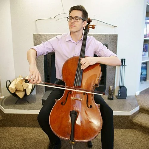 A man sitting in front of a fireplace playing a cello with a bow in a living room.