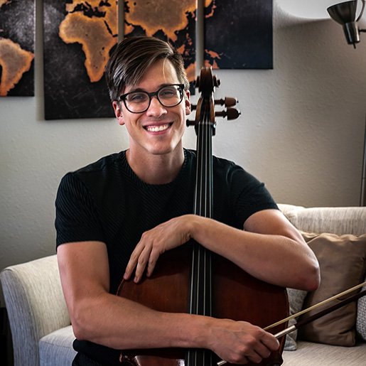 Young man with glasses smiling and holding a cello in a living room.