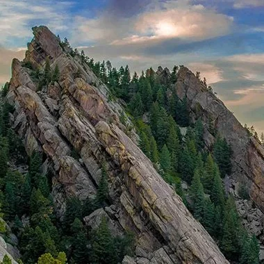 Steep rocky mountain slope with green trees and a partly cloudy sky.