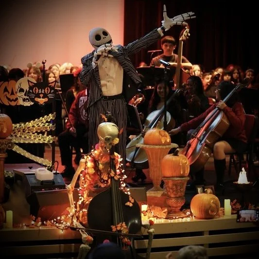 A Halloween-themed concert with a skeleton conductor leading a children's orchestra. The stage is decorated with pumpkins, skeletons, and Halloween decorations, and the children are playing string instruments.
