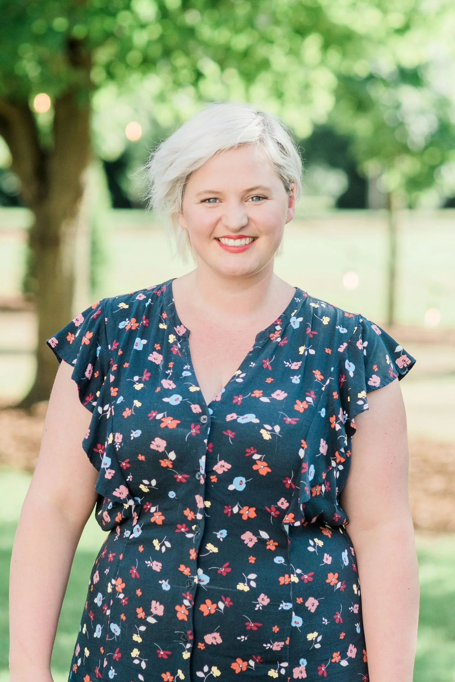 A smiling woman with short platinum blonde hair, wearing a navy floral dress, standing outdoors in a park with trees and greenery in the background.
